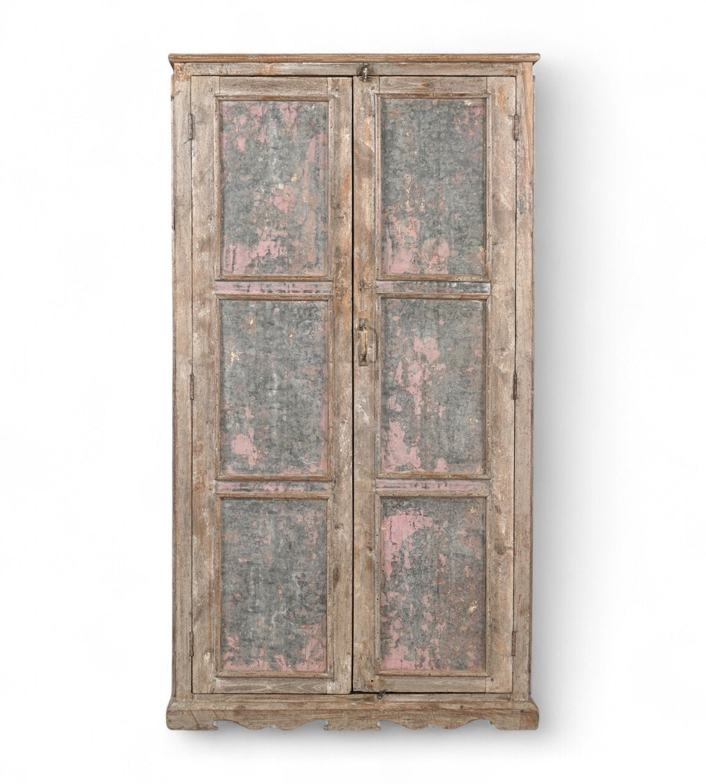 A vintage teak wood wardrobe with iron panels, featuring a carved base and an aged patina finish in soft pink and faded grey.