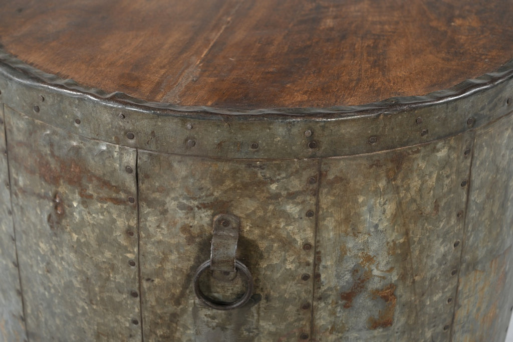 A cylindrical vintage side table made of aged iron with a teak wood top, featuring ring handles and a weathered finish.