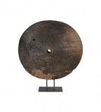 A large circular teak wood wheel with visible cracks and grooves, mounted on a metal iron stand.