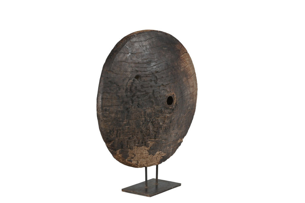 A large circular teak wood wheel with visible cracks and grooves, mounted on a metal iron stand.
